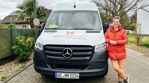 Eine Frau steht rechts neben einem Transporter. Auf dem Transporter ist die Aufschrift "Menschenmobil Herzogenrath e.V.". Die Frau lehnt sich an das Auto. Sie trägt eine rotorangene Jacke.