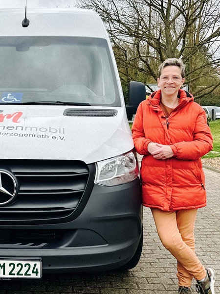 Eine Frau steht rechts neben einem Transporter. Auf dem Transporter ist die Aufschrift "Menschenmobil Herzogenrath e.V.". Die Frau lehnt sich an das Auto. Sie trägt eine rotorangene Jacke.