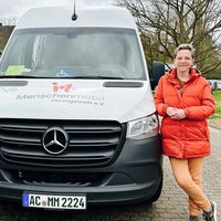 Eine Frau steht rechts neben einem Transporter. Auf dem Transporter ist die Aufschrift "Menschenmobil Herzogenrath e.V.". Die Frau lehnt sich an das Auto. Sie trägt eine rotorangene Jacke.