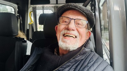 Ein älterer Mann lächelt in die Kamera. Er trägt eine Brille und eine Schiebermütze.