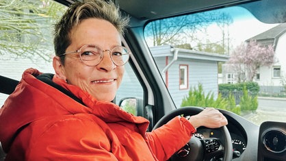 Eine Frau mit Brille sitzt am Steuer und dreht sich zur Kamera nach hinten rechts. Sie lächelt und trägt eine rotorangene Jacke.