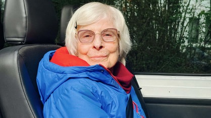 Eine ältere Frau mit Brille schaut in die Kamera. Sie lächelt und trägt eine blaue Jacke und darunter einen roten Pullover.