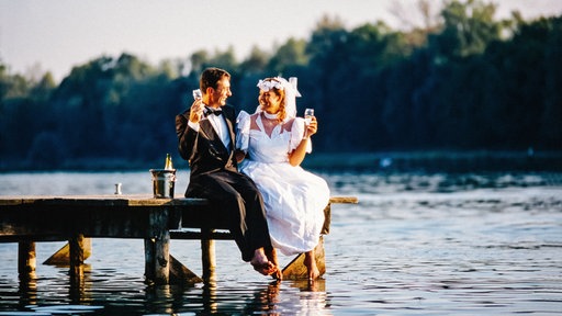 Ein Brautpaar sitzt auf einem Steg am Wasser und hält ein Sektglas in der Hand.