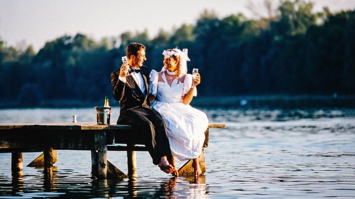 Ein Brautpaar sitzt auf einem Steg am Wasser und hält ein Sektglas in der Hand.