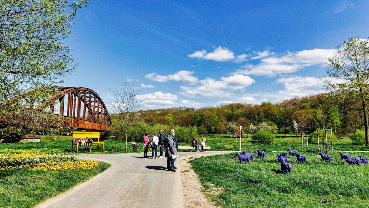 Besucher stehen auf einem Weg zwischen Wiesen und Beeten. Links im Bild befindet sich eine große Brücke. Auf einer Wiese rechts stehen blaue Schafs-Skulpturen.