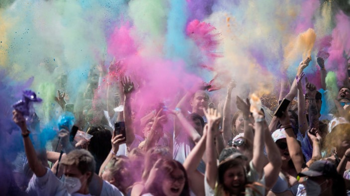 Menschen recken ihre Arme in die Luft und werfen Farbbeutel, um sie herum sind bunte Staubwolken zu sehen in Grün, Lila, Gelb und Blau.