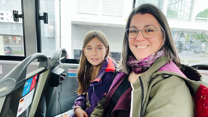 Christina Witte, eine Frau mittleren Alters, und ihre elfjährige Tochter Linda sitzen nebeneinander auf dem Oberdeck eines Doppeldeckerbusses und lächeln.