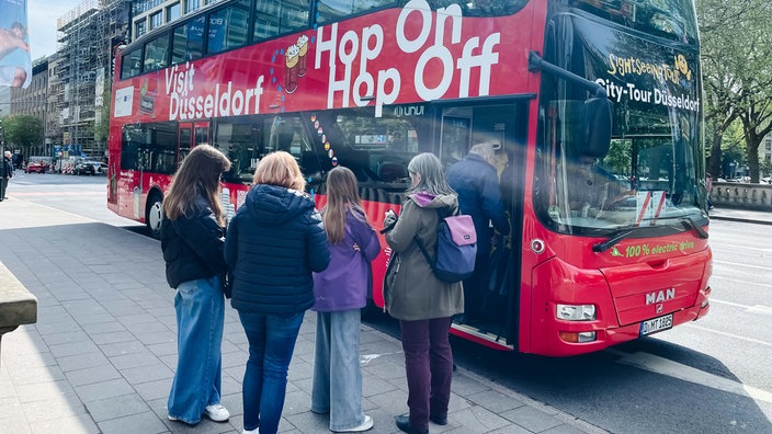 Ein roter Doppeldeckerbus steht in Düsseldorf an der Königsallee, davor warten Menschen.