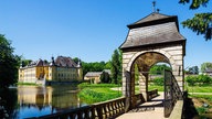 Eine Brücke mit einem Tor führt in den Garten von Wasserschloss Dyck, das sich im Hintergrund befindet.