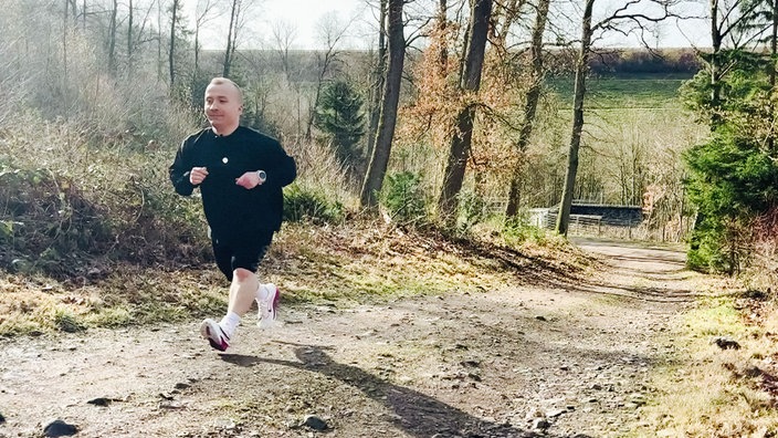 Maik Figgen, ein kleinwüchsiger braunhaariger Mann im schwarzen Trainingsanzug mit pinken Laufschuhe, läuft von der Kamera weg auf einem asphaltierten Weg durch den Wald.