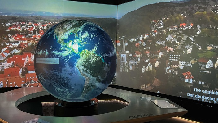 Ein Globus steht auf einem Podest, im Hintergrund ist eine Stadt auf eine Leinwand projiziert 