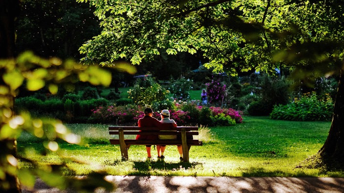 Ein Paar sitzt entspannt Arm in Arm auf einer Parkbank in einem blühenden Garten.