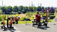 Ein Mann sitzt auf einem Sportgerät im Neuland-Park in Leverkusen. Im Hintergrund befindne sich Schornsteine.