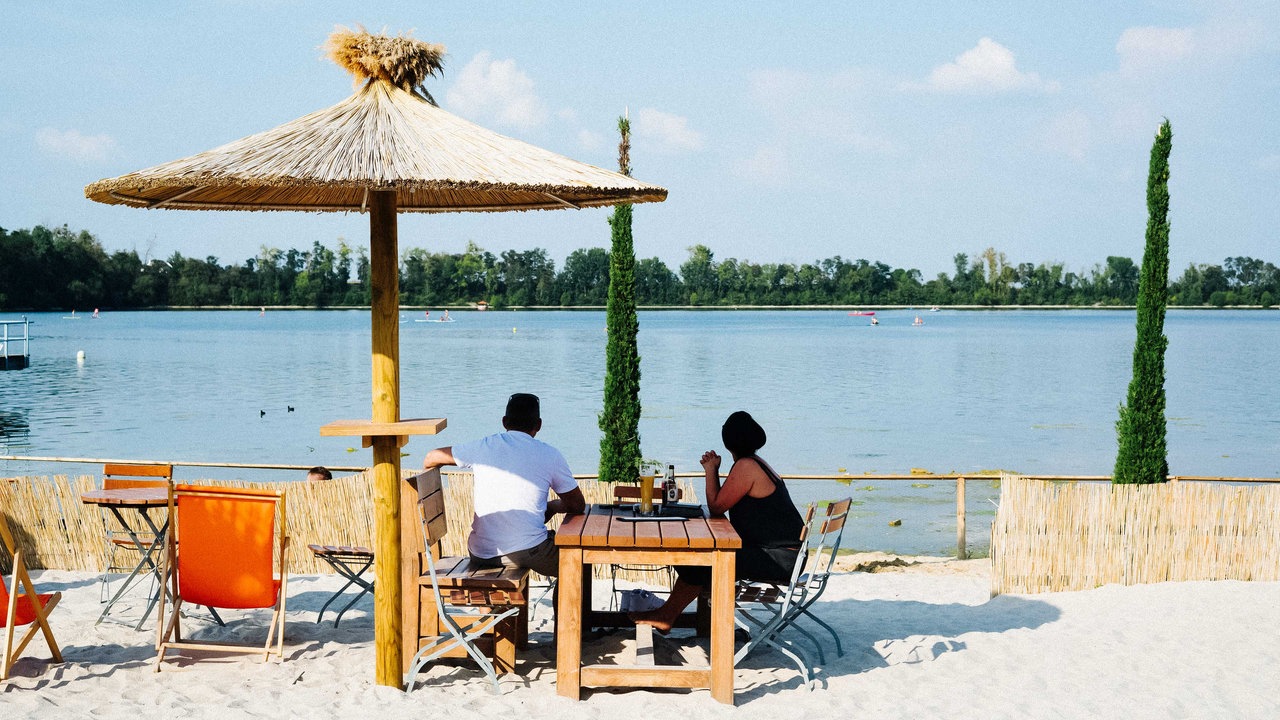 Ein Mann und eine Frau sitzen unter einem Sonnenschirm an einem Tisch und blicken auf den See.