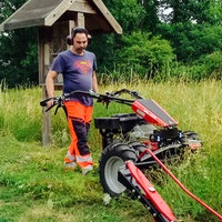 Ein Mann mäht mit einer Mähmaschine eine Wiese, um ihn herum liegen bereits geschnittene Halme
