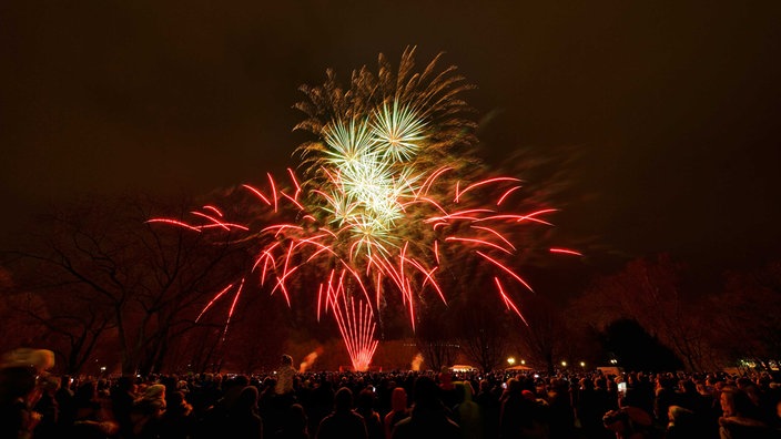 Dunkle Schemen von Menschen, die ihre Köpfen gen Himmel recken und dort ein rot-gelb-orangenes Feuerwerk sehen