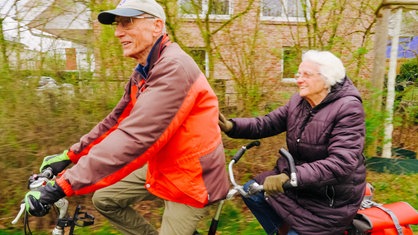 Ein älteres Ehepaar fährt gemeinsam Tandem. Die Frau hinten klopft dem Mann, der vorne fährt, auf den Rücken. 