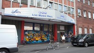Die Live Music Hall bei Tag von außen. Über dem Eingang steht "Live Music Hall", davor warten einige ältere Menschen in der Schlange. Links neben dem Eingang sind Graffiti zu sehen.