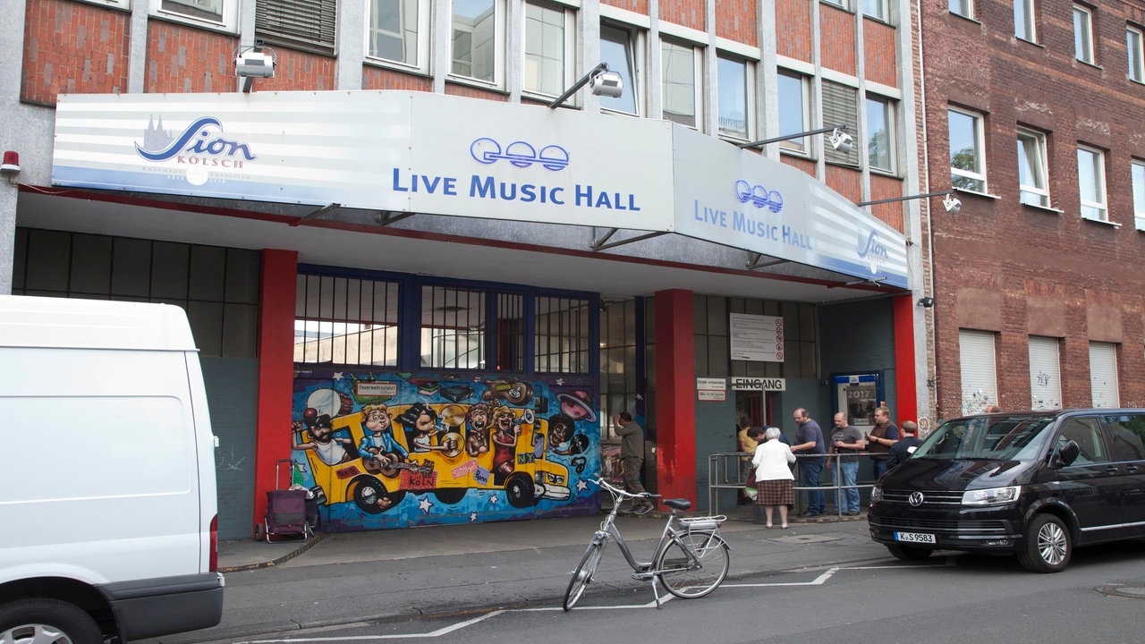 Die Live Music Hall bei Tag von außen. Über dem Eingang steht "Live Music Hall", davor warten einige ältere Menschen in der Schlange. Links neben dem Eingang sind Graffiti zu sehen.