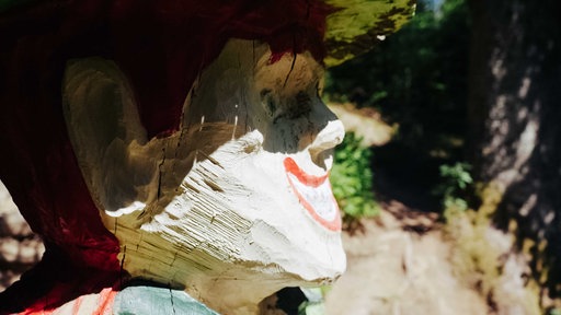 Seitenansicht auf eine aus Holz geschnitze Kobold-Figur mit roten Haaren, großen Ohren und einem rot umrandeten Lächeln.