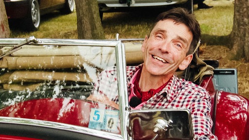 Markus Grünhagen sitzt in einem rot-blau karierten Hemd in einem roten Oldtimer und lächelt.