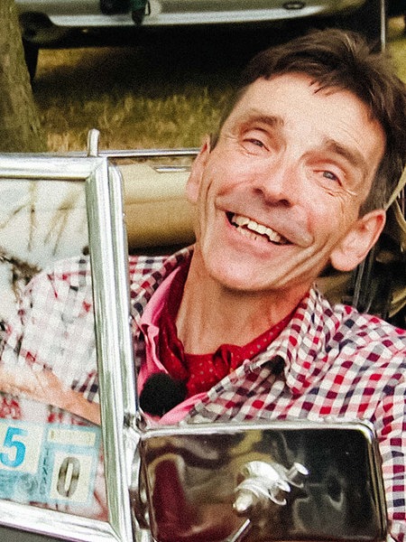 Markus Grünhagen sitzt in einem rot-blau karierten Hemd in einem roten Oldtimer und lächelt.