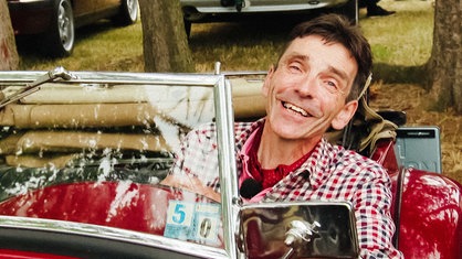 Markus Grünhagen sitzt in einem rot-blau karierten Hemd in einem roten Oldtimer und lächelt.
