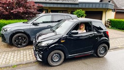 Johanna König sitzt in ihrem Mopedauto, das deutlich kleiner ist, als das dahinter geparkte, normale Auto.