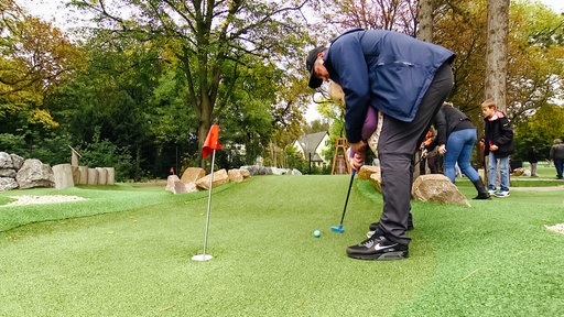 Ein Mann mit blauer Jacke, schwarzen Schuhen und schwarzer Hose hält einen Golfschläger.