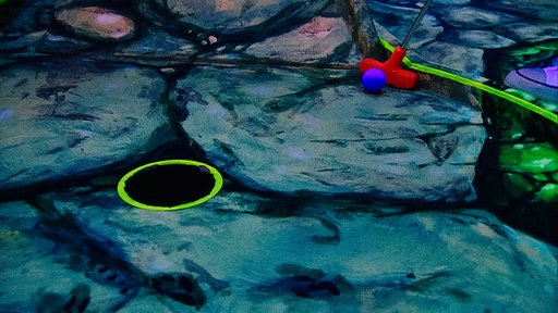 Ein Loch mit gelbem Neon-Rand auf Steinen, dahinter sieht man einen lila Golfball, der gerade von einem roten Golfschläger geschlagen wird. 