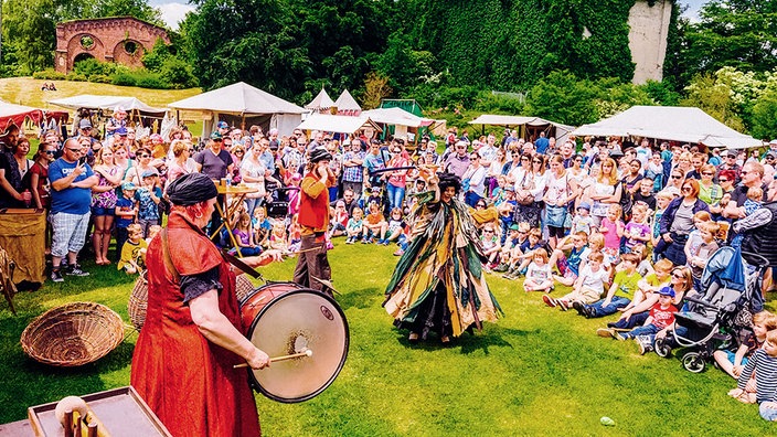 Eine Gruppe in mittelelterlicher Kleidung musiziert und tanzt auf einer Wiese. Ringsherum stehen und sitzen viele Erwachsene und Kinder und schauen zu. Im Hintergrund befinden sich verschiedene Stände.