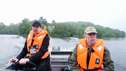 Johannes Pantel und Michael Knappstein im Motorboot im Wasser, einer am Lenkrad, der andere sitzt daneben, beide tragen orange Rettungswesten.