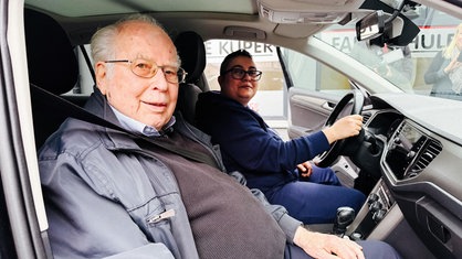Wolfgang Küper sitzt neben seiner Schülerin im Fahrschulauto mit offenen Türen. Beide schauen in die Kamera.