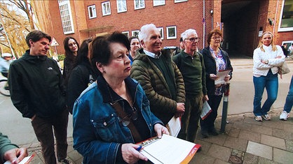 Eine Gruppe von Menschen verschiedenen Alters steht auf der Straße vor einem Backsteinhaus und hält Fragebögen in der Hand.