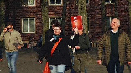 Vier Personen vor einem roten Backsteingebäude folgen Vera Isabell Blasum, einer rothaarigen Frau im schwarzen Mantel, die rote Karten hochhält.