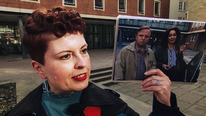 Vera Isabell Blasum, eine rothaarige Frau im schwarzen Mantel, hält mit der linken Hand ein Foto von zwei Personen hoch, den Tatort-Darstellern Axel Prahl und Mechthild Großmann.