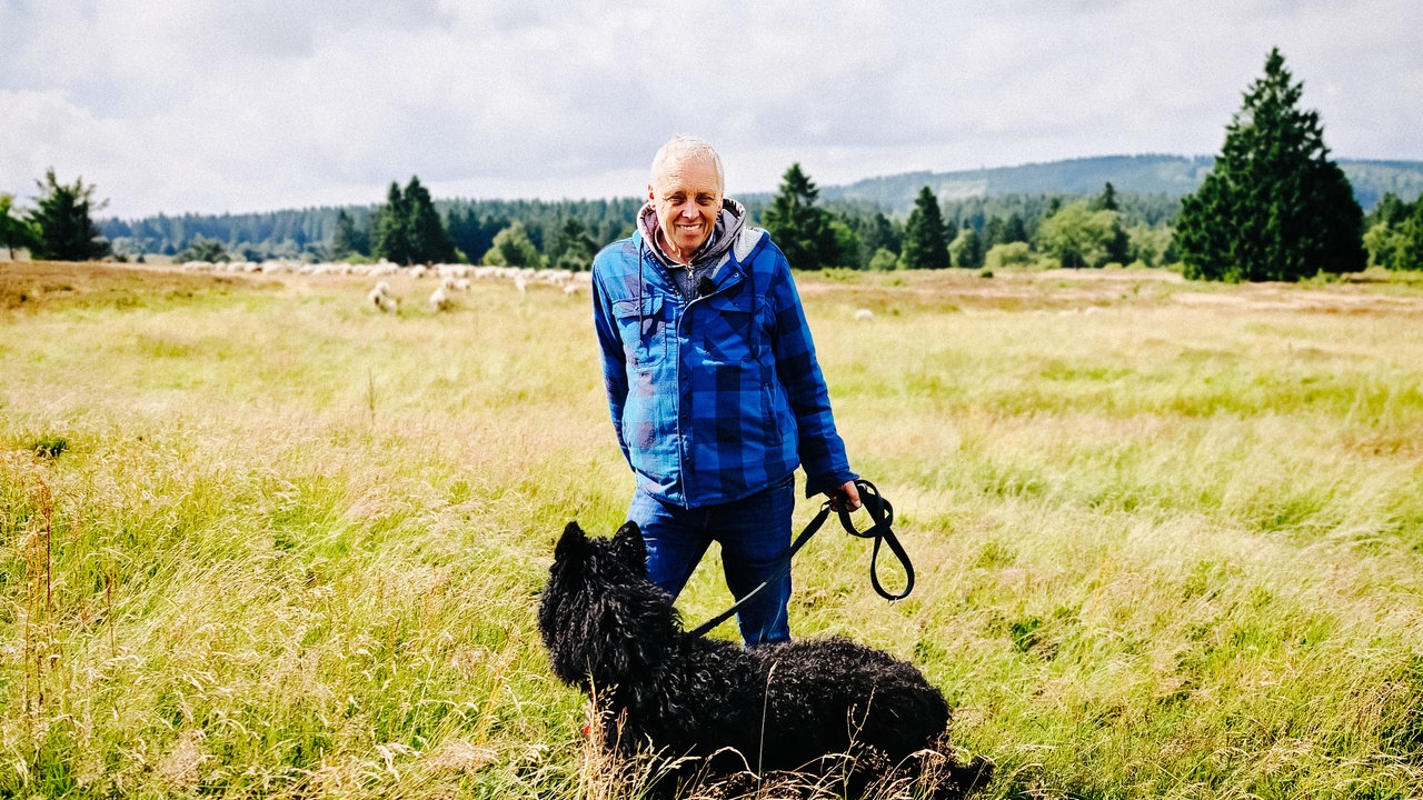 Schäfer Peter Kuhlmann steht mit seinem Hund auf der Hochheide. Im Hintergrund sind seine Schafe.