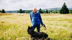 Schäfer Peter Kuhlmann steht mit seinem Hund auf der Hochheide. Im Hintergrund sind seine Schafe.