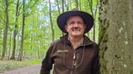 Eifel-Ranger Ralf Hilgers lehnt mit braunem Hut und brauner Jacke an einem Baum im Nationalpark Eifel