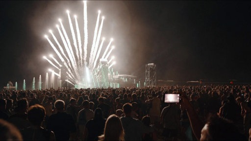 Eine Menschenmenge steht vor einer großen Festivalbühne, über der Feuerwerk explodiert.