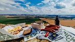 Ein Picknick mit Brot, Käse, Würsten liegt auf einer Steinfläche vor einer weiten, hügeligen Landschaft mit Feldern.