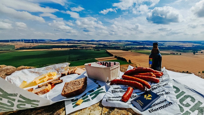 Ein Picknick mit Brot, Käse, Würsten liegt auf einer Steinfläche vor einer weiten, hügeligen Landschaft mit Feldern.