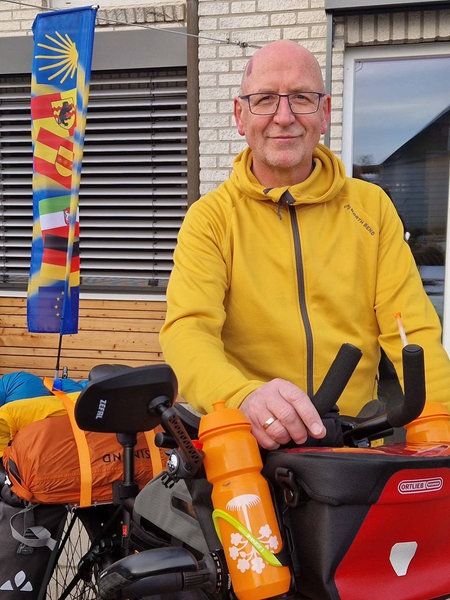 Thomas Fischer steht in einer gelben Jacke neben seinem voll bepackten und mit einer großen Fahne geschmückten Fahrrad.
