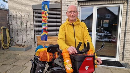 Thomas Fischer steht in einer gelben Jacke neben seinem voll bepackten und mit einer großen Fahne geschmückten Fahrrad.