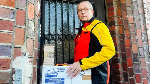 Ein Mann lehnt mit einer DHL Paketboten Jacke vor einem Hauseingang und hält zwei Pakete aus Pappe in der Hand.