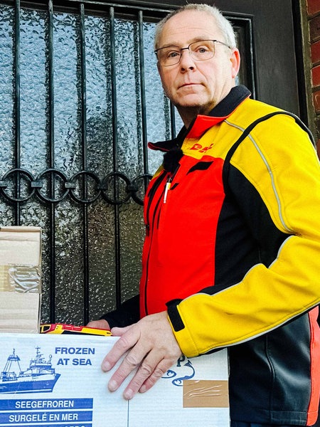 Ein Mann lehnt mit einer DHL Paketboten Jacke vor einem Hauseingang und hält zwei Pakete aus Pappe in der Hand.