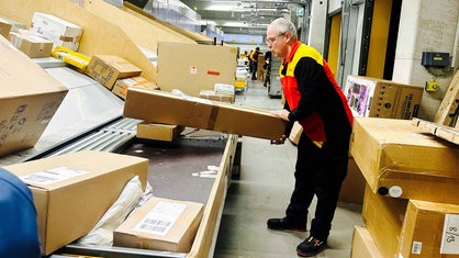 Ein Mann steht in einer Lagerhalle mit vielen Paketen in verschiedenen Größen und hält einen großen Karton in der Hand. Er trägt eine Weste mit dem Logo von DHL