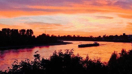 Sonnenuntergang über einem großen Fluss, auf dem eine kleine Fähre fährt. 