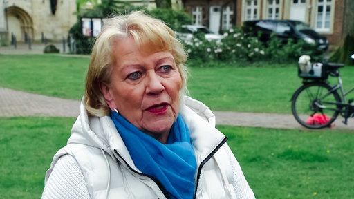Frau mit blinen kurzen Haaren, blauem Schal und weißer Jacke, Blick nach vorne gerichtet, den Mund leicht geöffnet, hinter ihr Wiese und ein Weg