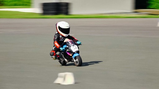 Kleiner Junge auf einem Mini-Motorrad auf einer Rennstrecke, er trägt einen weißen Helm und schwarz-rote Schutzkleidung mit blauen Handschuhen.